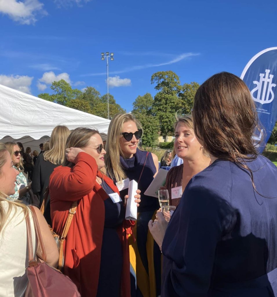 Skolans alumner minglar i strålande solsken på idrottsplanen vid sjön. 