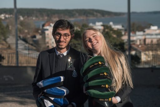 Två glada elever som bär på högar av mössor - 50 dagar kvar till studenten!
